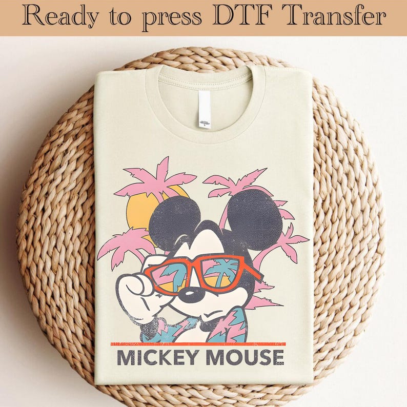 May include: A beige t-shirt with a vintage-style graphic of Mickey Mouse wearing sunglasses and a Hawaiian shirt. The graphic is set against a background of pink palm trees and a yellow sun. The text "MICKEY MOUSE" is printed below the graphic.
