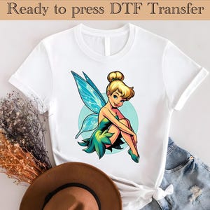 Puede incluir: Camiseta blanca con una imagen de dibujos animados de color de Campanilla, un hada con alas verdes y un vestido verde, sentada en un círculo azul.