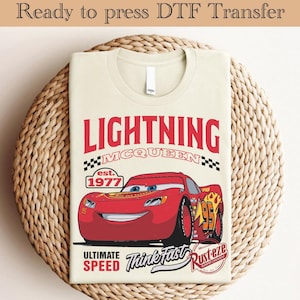 Può includere: Una maglietta beige con una grafica rossa, bianca e nera di Saetta McQueen dal film Cars. La grafica include il testo "Lightning McQueen", "est. 1977", "Ultimate Speed", "Think Fast" e "Rust-eze", insieme a un motivo a scacchi.