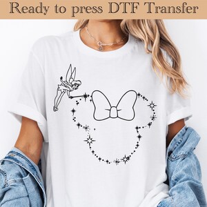 May include: White t-shirt with a black and white design of Tinkerbell sprinkling fairy dust on a bow. The design is in the shape of Mickey Mouse ears.
