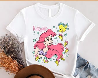 May include: White t-shirt featuring a colorful graphic of Ariel from The Little Mermaid. The design includes Ariel with pink hair, a purple seashell bra, and yellow seahorse-like creatures. The text "The Little Mermaid" is also visible.