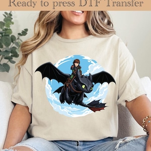 May include: Beige t-shirt featuring a cartoon graphic of a black dragon with a rider against a blue sky background. The text "Ready to press DTF Transfer" is at the top. The design is centered on the shirt, offering a playful and adventurous theme.