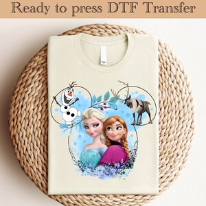 May include: A cream colored t-shirt with a printed design featuring Elsa and Anna from Frozen, Olaf the snowman, and a reindeer. The design is in a Mickey Mouse shape with blue, white, and green accents.