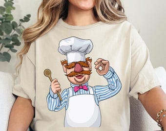 Pode incluir: Camiseta bege com um chef de desenho animado com chapéu branco, bigode grande e gravata borboleta rosa. O chef segura uma colher de pau e faz um gesto de mão 'ok'. O texto na parte superior diz 'Ready to press DTF Transfer'.