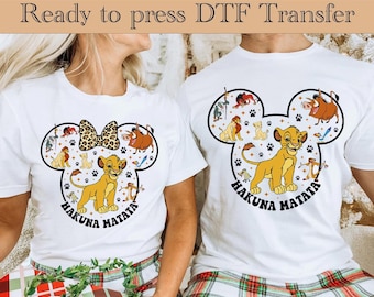 Könnte beinhalten: Weißes T-Shirt mit einem Mickey-Maus-Kopf-Design mit Figuren aus dem Film Der König der Löwen. Das Design beinhaltet Simba, Pumbaa, Timon und andere Figuren. Der Text "Hakuna Matata" ist unter dem Design gedruckt.