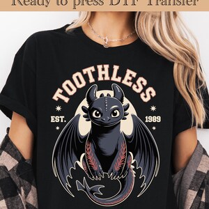 May include: Black t-shirt featuring a cartoon dragon with the text "TOOTHLESS" above the dragon and "EST. 1989" below. The dragon is black with red accents on its wings and is set against a cream-colored circle.