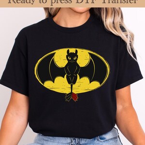 May include: Black t-shirt featuring a cartoon dragon design within a yellow oval. The dragon is black with yellow eyes and a red and black tail. The text "Ready to press DTF Transfer" is at the top.