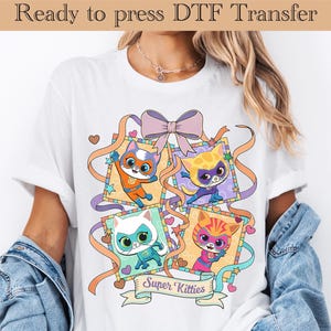 Könnte beinhalten: Weißes T-Shirt mit einem farbenfrohen Design von vier Superhelden-Katzen. Das Design beinhaltet eine Schleife, Bänder und den Text "Super Kitties". Der obere Text lautet "Ready to press DTF Transfer".