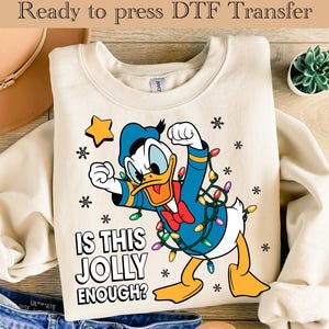 May include: A cream-coloured sweatshirt with a Donald Duck graphic in a festive design. Donald is wrapped in Christmas lights, with snowflakes and a star. The text reads "IS THIS JOLLY ENOUGH?" A DTF transfer for the holidays.