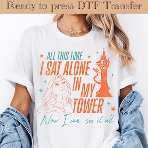 May include: White t-shirt with a graphic design featuring the text "ALL THIS TIME I SAT ALONE IN MY TOWER" and an illustration of a character looking up at a tower. The design is in orange and teal.