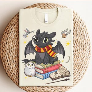 May include: A light beige t-shirt with a cartoon dragon wearing a scarf and glasses, sitting on books. The design includes a golden snitch, a key, and an owl. The shirt is folded. The design is inspired by a popular fantasy series.