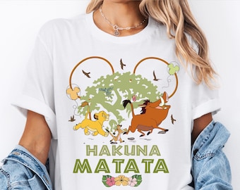 Könnte beinhalten: Weißes T-Shirt mit einem farbenfrohen Aufdruck von Charakteren aus Der König der Löwen, darunter Simba, Timon und Pumbaa. Das Design enthält die Worte "HAKUNA MATATA" in Grün mit floralen Akzenten. Der Text "Ready to press DTF Transfer" steht oben.