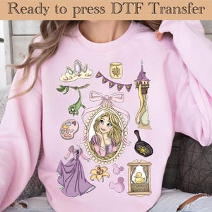 Op de afbeelding: Roze sweatshirt met een DTF-transferontwerp met verschillende cartoon-elementen. Het ontwerp bevat een prinsessenportret, een toren, een koekenpan en andere gerelateerde afbeeldingen. De tekst "Ready to press DTF Transfer" staat bovenaan.