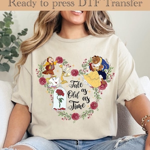 May include: Beige t-shirt featuring a heart-shaped design with characters from Beauty and the Beast. The design includes Belle in a yellow dress, the Beast, and the text "Tale as Old as Time".