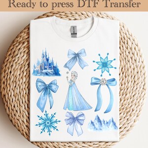 May include: A white t-shirt with a blue and white design featuring a castle, a snowflake, a princess, and bows. The design is ready to be pressed using a DTF transfer.