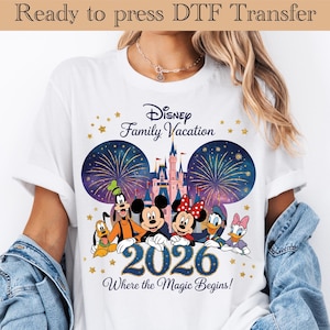 Op de afbeelding: Wit T-shirt met een Disney Family Vacation ontwerp. De afbeelding toont Mickey en Minnie Mouse, Donald en Daisy Duck, Goofy en Pluto, met een kasteel en vuurwerk. Het jaar 2026 staat afgebeeld, met de tekst "Where the Magic Begins!"