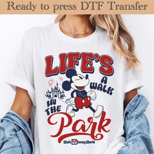 May include: White t-shirt featuring Mickey Mouse and the text "LIFE'S A WALK IN THE PARK" in red and blue. The design includes a castle, fireworks, and the Walt Disney World logo. The shirt is ready to press.