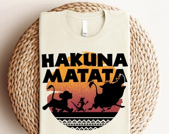 Könnte beinhalten: Ein beiges T-Shirt mit einem schwarz-weißen Grafikdruck des Satzes "Hakuna Matata" mit einem Sonnenuntergangs-Hintergrund und Silhouetten von drei Tieren.