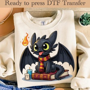 May include: Cream-colored sweatshirt with a cartoon dragon graphic. The black dragon wears glasses and a scarf, sitting on books with candles and a flame. Text at the top reads "Ready to press DTF Transfer."