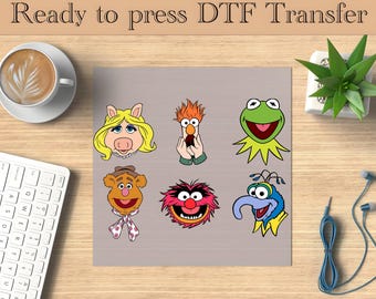 Puede incluir: Una hoja con caras de personajes de dibujos animados, incluyendo Miss Piggy, Beaker, Kermit la Rana, Fozzie Bear, Animal y Gonzo. El texto "Ready to press DTF Transfer" está en la parte superior. La imagen está sobre una superficie de madera con una taza de café, un teclado y una planta.
