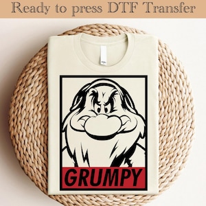 以下が含まれることがあります： 白雪姫のグランピーの白黒漫画イラストが描かれたクリーム色のTシャツ。「GRUMPY」の文字がデザインの下部に黒い長方形に赤色でプリントされています。「Ready to press DTF Transfer」の文字が上部にあります。