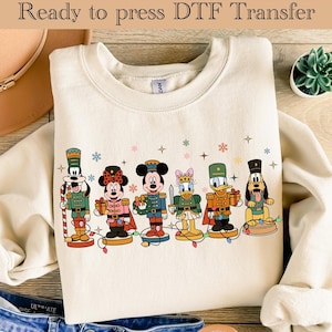 May include: Cream-colored sweatshirt with a festive design featuring cartoon characters dressed as nutcrackers. The characters include Mickey Mouse, Minnie Mouse, Donald Duck, and Goofy, with the text "Ready to press DTF Transfer" at the top.