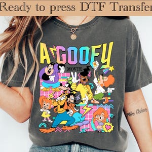 Puede incluir: Una camiseta gris carbón con un gráfico colorido de "A Goofy Movie". El diseño incluye varios personajes de dibujos animados en colores vibrantes, con el título de la película en una fuente audaz y multicolor. Las palabras "Ready to press DTF Transfer" están en la parte superior.