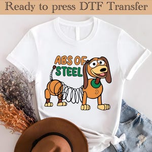 Può includere: Maglietta bianca con un'immagine di Slinky Dog di Toy Story in stile cartone animato. Il testo "ABS OF STEEL" è stampato sopra l'immagine.