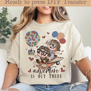 May include: Beige t-shirt with a colorful graphic featuring two cartoon characters in aviator gear on a plane, with balloons and a house. The text "adventure is out there" is below the image. The words "Ready to press DTF Transfer" are at the top.
