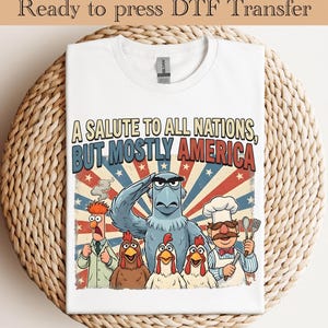 May include: White t-shirt with a graphic featuring cartoon characters and the text "A SALUTE TO ALL NATIONS, BUT MOSTLY AMERICA." The design includes an eagle, chickens, and other characters in a patriotic theme.