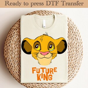 May include: A beige t-shirt with a cartoon image of Simba from The Lion King. The image is in the center of the shirt and is surrounded by the text "FUTURE KING" in orange.