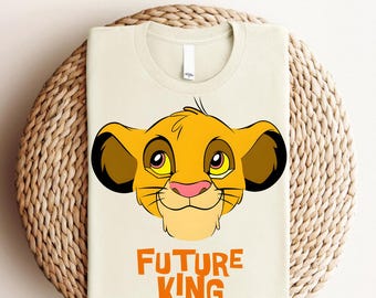 Könnte beinhalten: Ein beiges T-Shirt mit einem Cartoon-Bild von Simba aus dem König der Löwen. Das Bild befindet sich in der Mitte des Shirts und ist umgeben vom Text "FUTURE KING" in Orange.