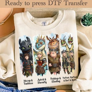 May include: Cream-colored sweatshirt featuring a DTF transfer with characters from the "How to Train Your Dragon" franchise. The design includes Hiccup & Toothless, Astrid & Stormfly, Fishlegs & Meatlug, and Tuffnut, Ruffnut & Barf & Belch. Text at the top reads "Ready to press DTF Transfer."