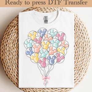 Op de afbeelding: Wit T-shirt met een ontwerp van Mickey Mouse-vormige ballonnen in pasteltinten. Onderaan een roze strik. De tekst "Ready to press DTF Transfer" staat bovenaan. Het T-shirt ligt op een geweven oppervlak.