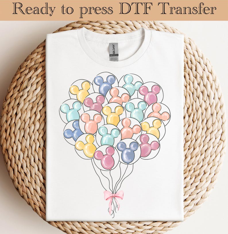 Op de afbeelding: Wit T-shirt met een ontwerp van Mickey Mouse-vormige ballonnen in pasteltinten. Onderaan een roze strik. De tekst "Ready to press DTF Transfer" staat bovenaan. Het T-shirt ligt op een geweven oppervlak.