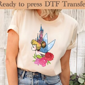 May include: A white t-shirt with a graphic print of Tinkerbell flying in front of a pink castle with flowers. The text "Ready to press DTF Transfer" is at the top of the image.