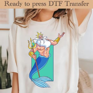 Könnte beinhalten: Ein weißes T-Shirt mit einem Cartoon-Bild eines blau-häutigen, bärtigen Mannes mit einer goldenen Krone und einem Dreizack. Der Mann hält den Dreizack in seiner rechten Hand und hat seine linke Hand erhoben. Der Hintergrund ist hellgrün.