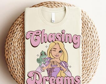 Pode incluir: Uma camiseta de cor creme com um gráfico rosa de uma princesa de desenho animado com cabelo longo e loiro e um lagarto verde. O gráfico diz "Chasing Dreams" em uma fonte retrô.