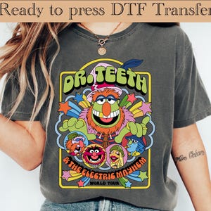 Op de afbeelding: Donkergrijs t-shirt met een kleurrijke afbeelding van Dr. Teeth and The Electric Mayhem World Tour. Het ontwerp bevat de bandnaam in groen en geel, met cartoonfiguren en sterren.