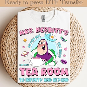 May include: White t-shirt featuring a cartoon image of Buzz Lightyear as Mrs. Nesbitt, with the text "MRS. NESBITT'S TEA ROOM TO INFINITY AND BEYOND." The design includes a teapot, hat, and other cartoon elements. Ready to press DTF transfer.