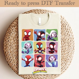 May include: A cream-coloured t-shirt with a colourful graphic of nine Spidey characters in square frames. The text "SPIDEY AMAZING FRIENDS" is below the characters. The shirt is displayed on a woven, circular surface. The text "Ready to press DTF Transfer" is at the top.