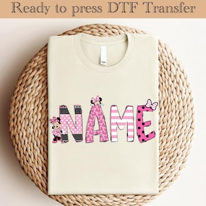 Pode incluir: Uma camiseta bege com um design rosa e preto da palavra "NAME" com personagens da Minnie Mouse.