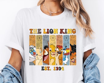 Könnte beinhalten: Weißes T-Shirt mit einer Retro-Grafik der König der Löwen-Charaktere, darunter Simba, Nala, Timon, Pumbaa, Zazu und Scar. Die Grafik ist von Feuerwerk umgeben und der Text "The Lion King Est. 1994".