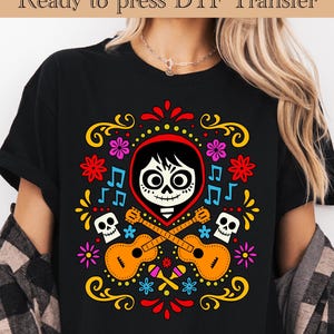 May include: Black t-shirt featuring a colorful graphic design inspired by the Day of the Dead. The design includes a skull, guitars, flowers, and musical notes. The text "Ready to press DTF Transfer" is at the top.