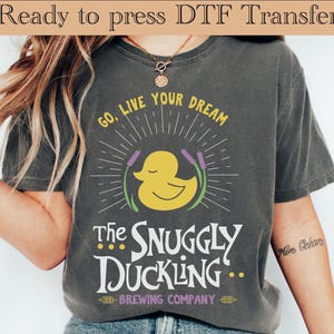 May include: Dark gray t-shirt featuring a yellow duck design with the text "Go, Live Your Dream" and "The Snuggly Duckling Brewing Company." The design includes a yellow duck, purple reeds, and radiating lines. The words "Ready to press DTF Transfer" are at the top.