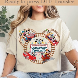 May include: Beige t-shirt featuring a colorful graphic of Mickey and Minnie Mouse on a roller coaster, with the text "Mickey & Minnie's Runaway Railway." The design includes a brown track and star accents. The words "Ready to press DTF Transfer" are at the top.