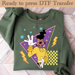 Puede incluir: Sudadera verde con una imagen de dibujos animados de un perro con una camisa amarilla sobre un fondo morado. El perro está en un diseño geométrico con rayos y un patrón a cuadros. El texto "Ready to press DTF Transfer" está en la parte superior de la imagen.