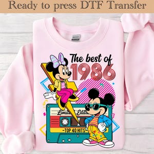 May include: A light pink sweatshirt featuring a colorful graphic of Minnie and Mickey Mouse. The design includes the text "The best of 1986" and a cassette tape. The sweatshirt is ready to press DTF transfer.