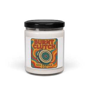 May include: A white soy candle in a glass jar with a black lid. The label features the text "BURNT CLUTCH" in orange, with a retro design of a clutch and the words "SOY CANDLE 9 OZ".
