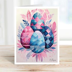 May include: Easter greeting card featuring four decorative eggs in shades of pink and blue, with geometric patterns. The eggs are surrounded by pink and blue leaves and feathers, creating a watercolor effect. The card is on a wooden surface.